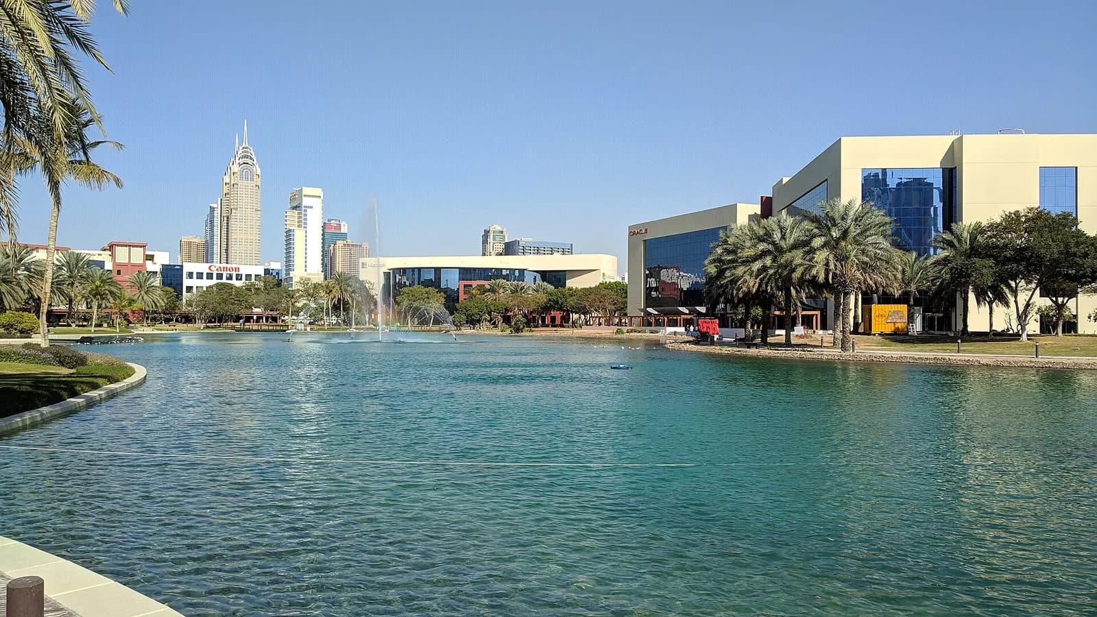Low-rise office buildings and palm trees around the lake at Dubai Internet City, with towers on Sheikh Zayed Road behind