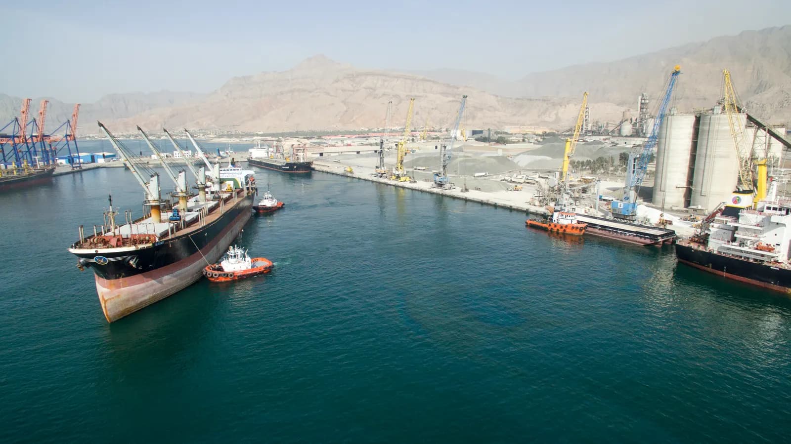 Bulk carriers and tugs at a Ras Al Khaimah port with cranes, silos and the Hajar mountains behind