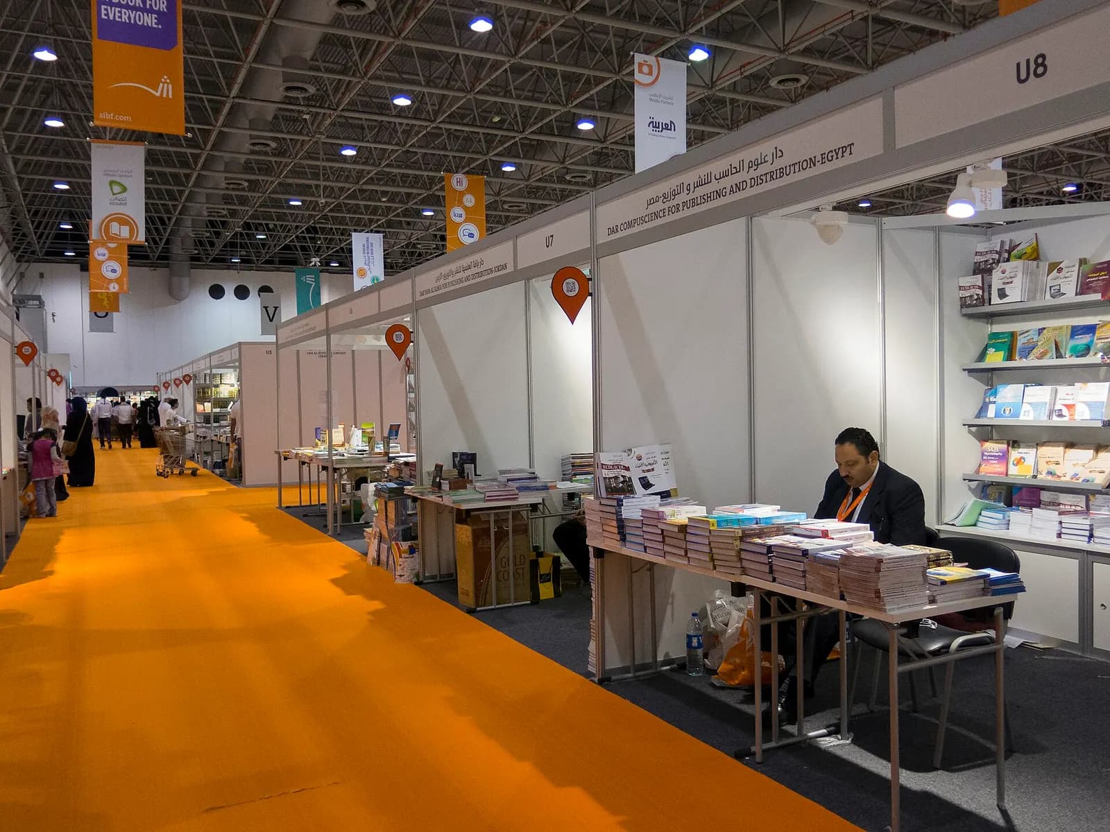An exhibitor seated at a book stand in a long hall of publishers' stalls at the Sharjah International Book Fair