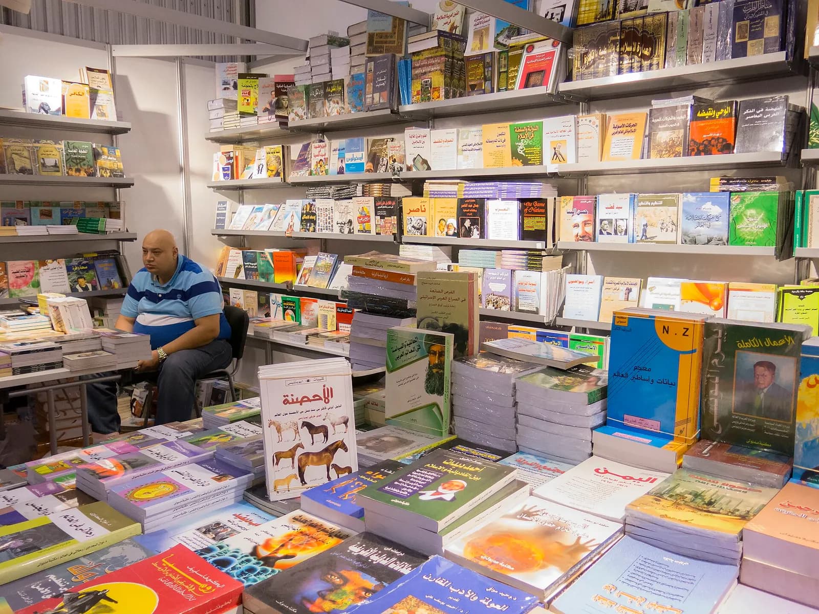 Shelves and tables of Arabic books at a publisher's stand, with a bookseller seated behind the display