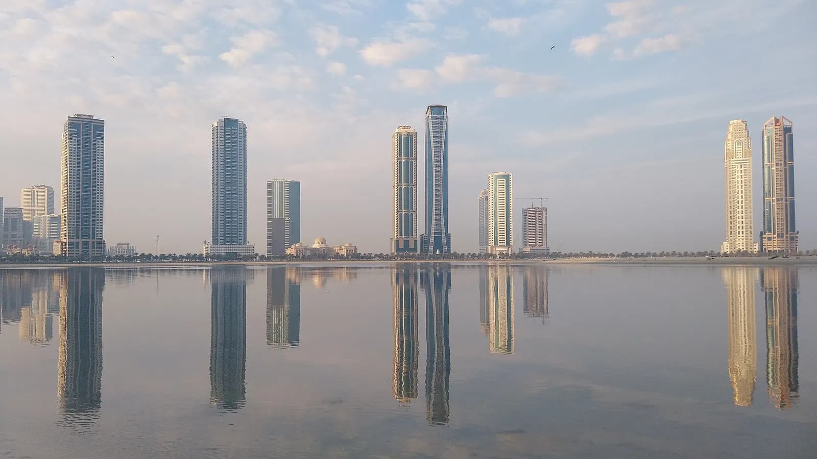 Residential and office towers reflected in calm water, seen from the Al Qasba corniche in Sharjah