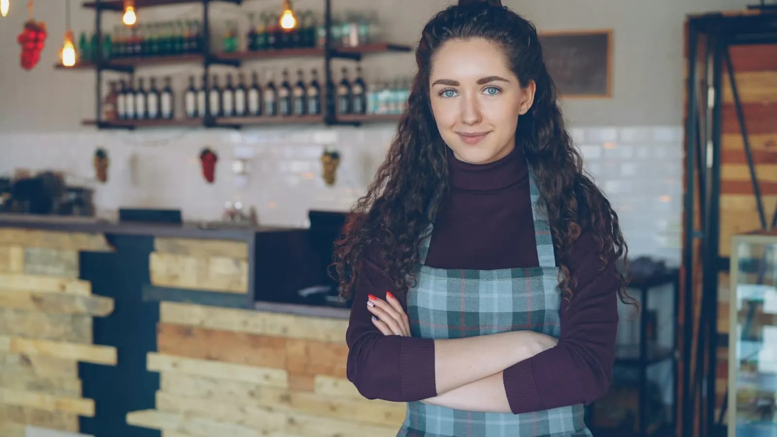 Young café owner in an apron standing with arms crossed behind her coffee shop counter