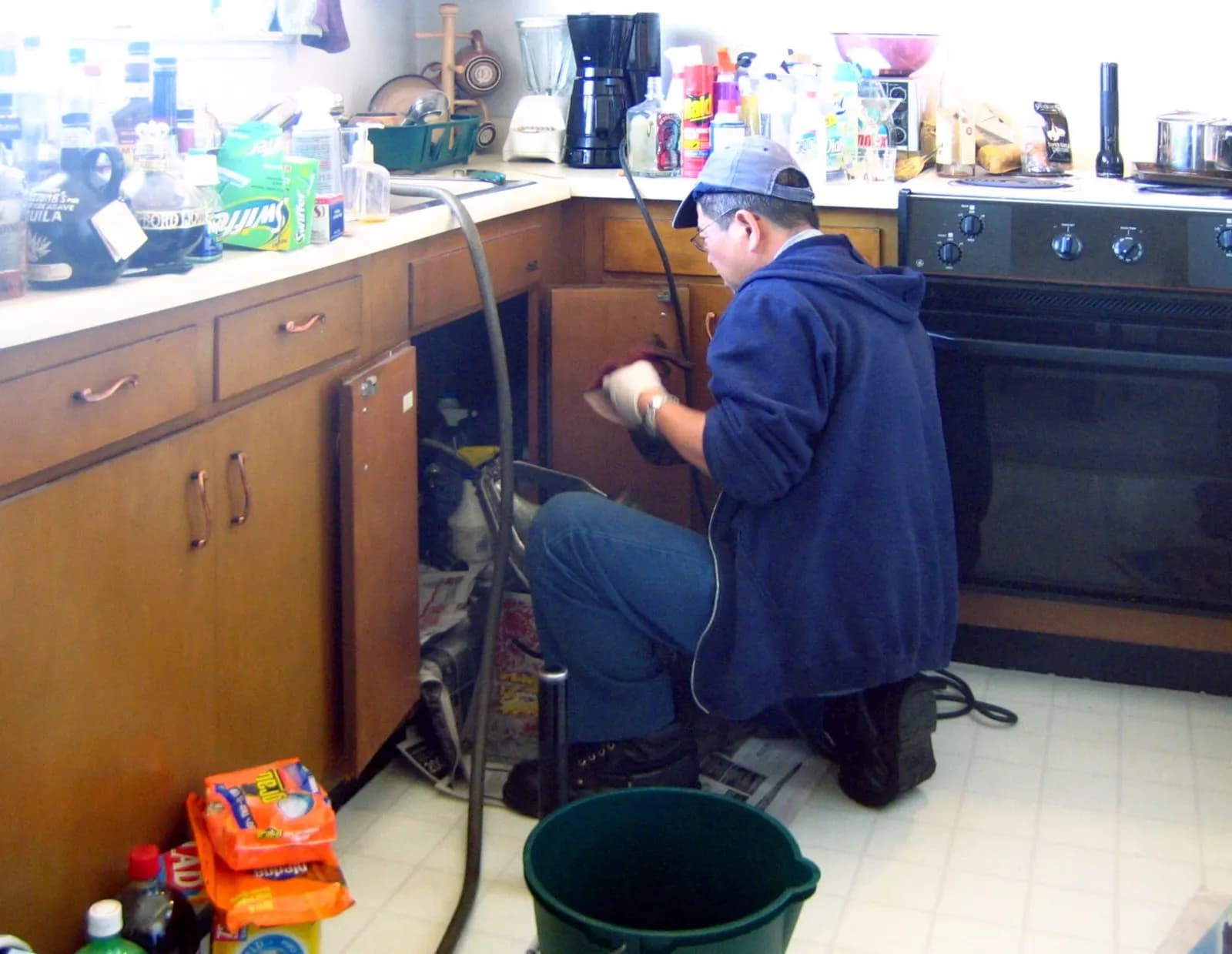 A plumber in a cap kneeling at an open kitchen cabinet, working on the pipes under the sink