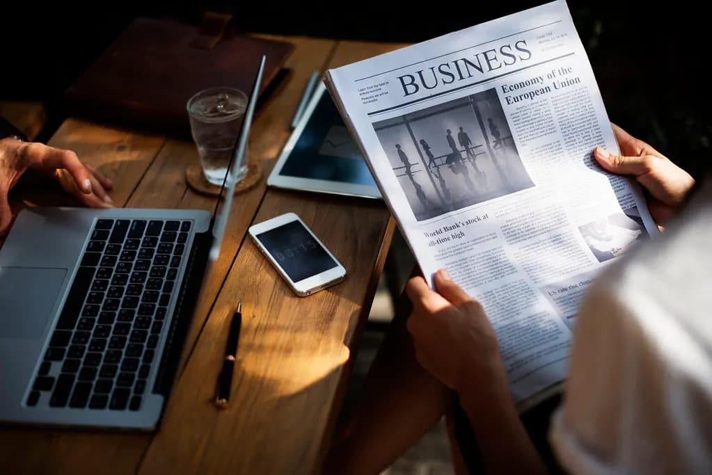 A laptop, phone and tablet on a wooden table beside a person reading a business newspaper