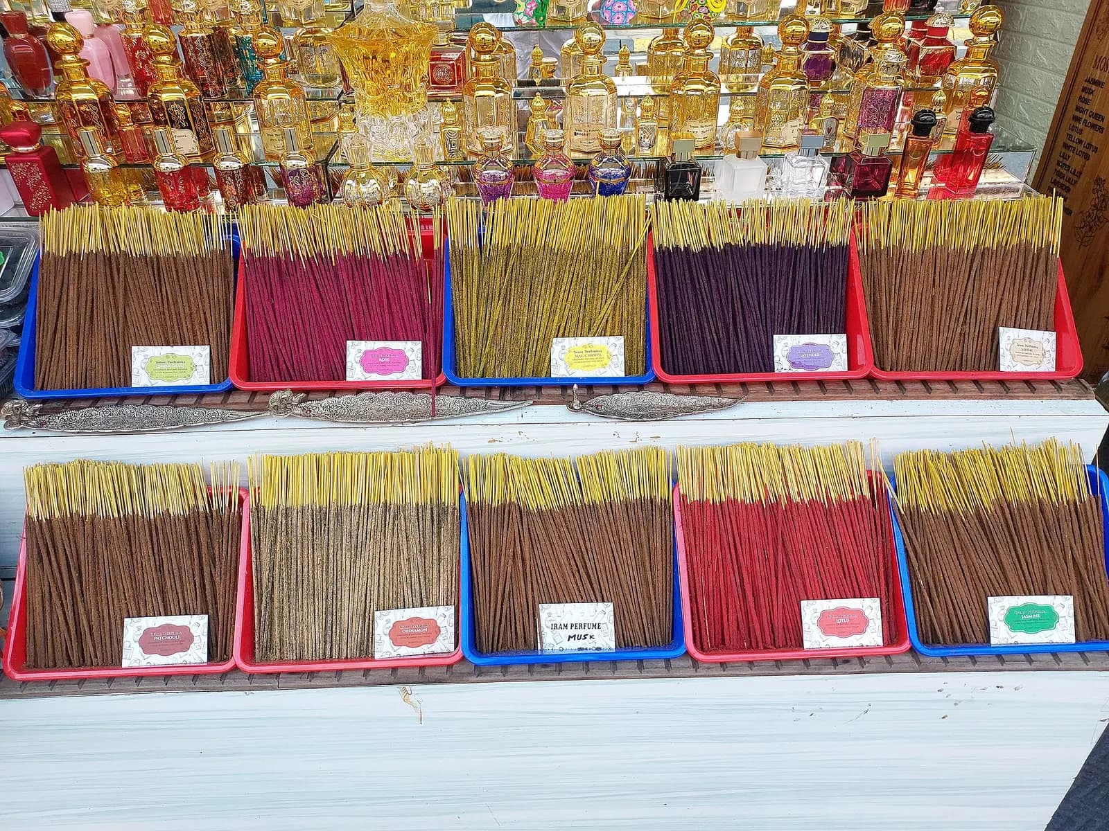 Loose scented agarbatti in coloured trays, sorted by fragrance, on a shop counter with perfume bottles behind