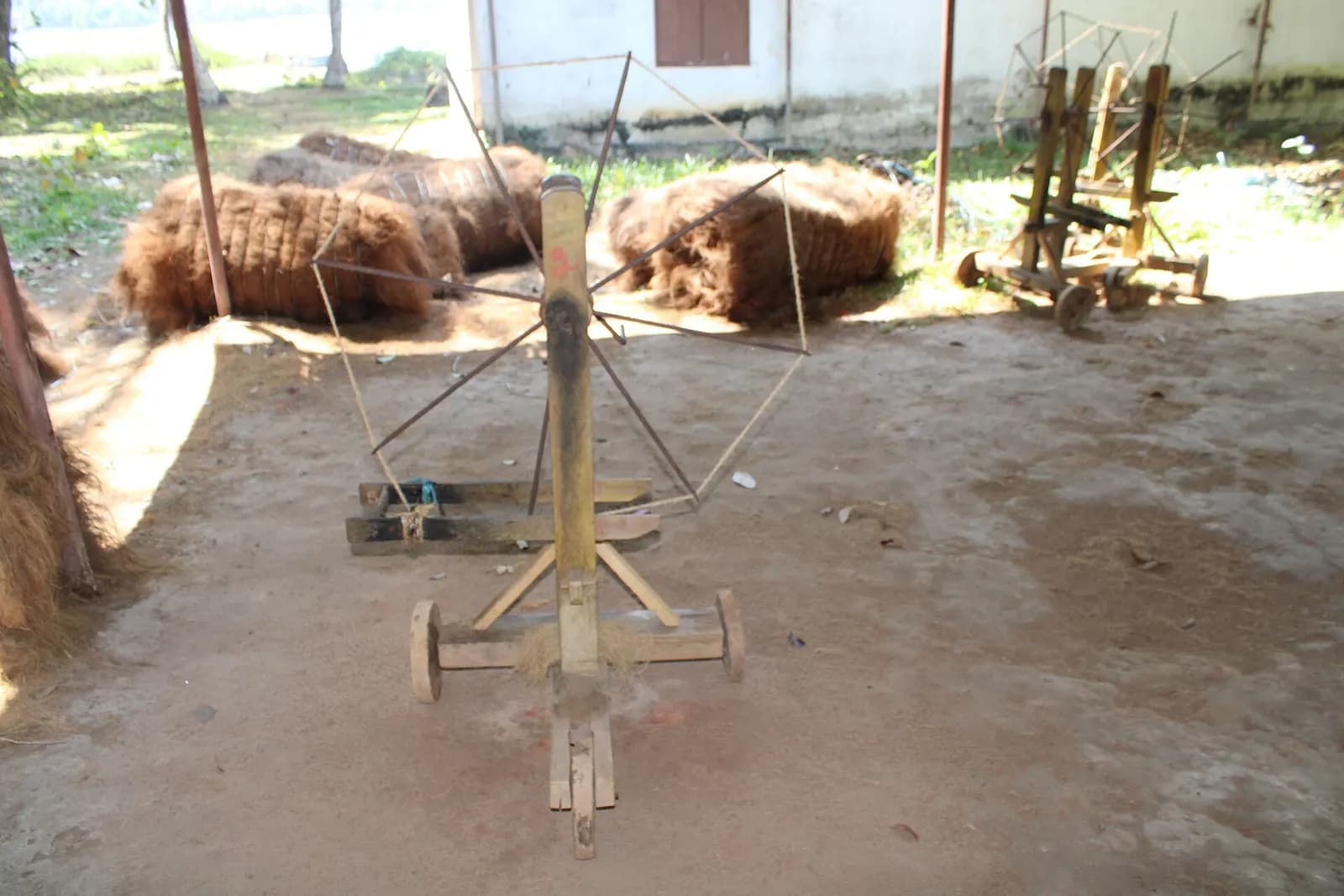 A simple coir spinning machine in a village yard with bundles of coconut husk fibre behind it