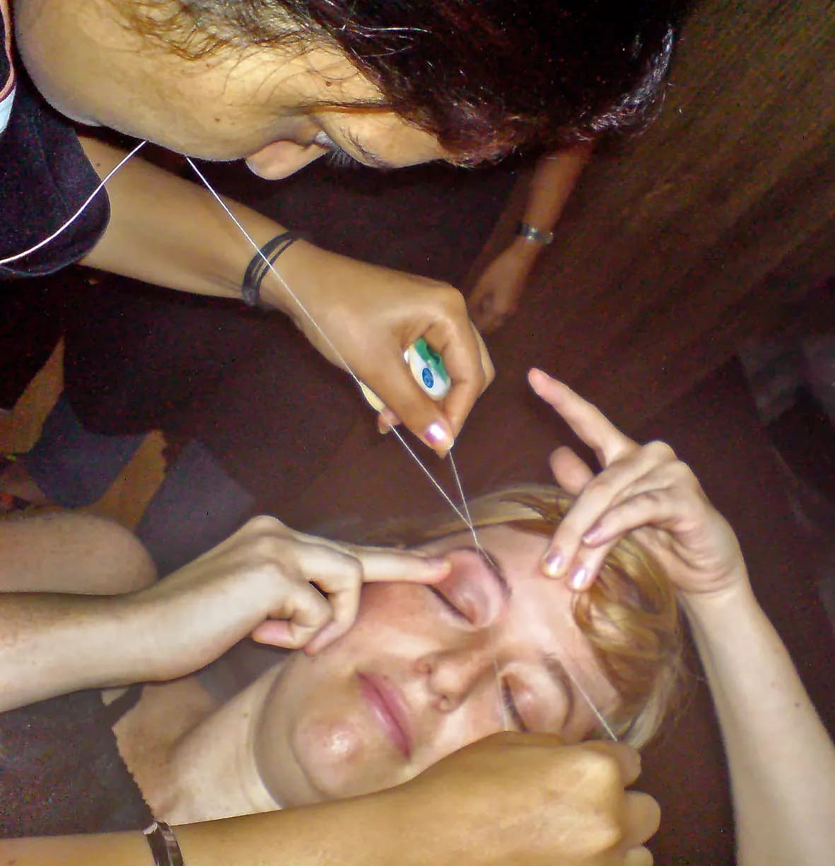 A beautician shaping a client's eyebrows with a cotton thread while the client lies back with eyes closed