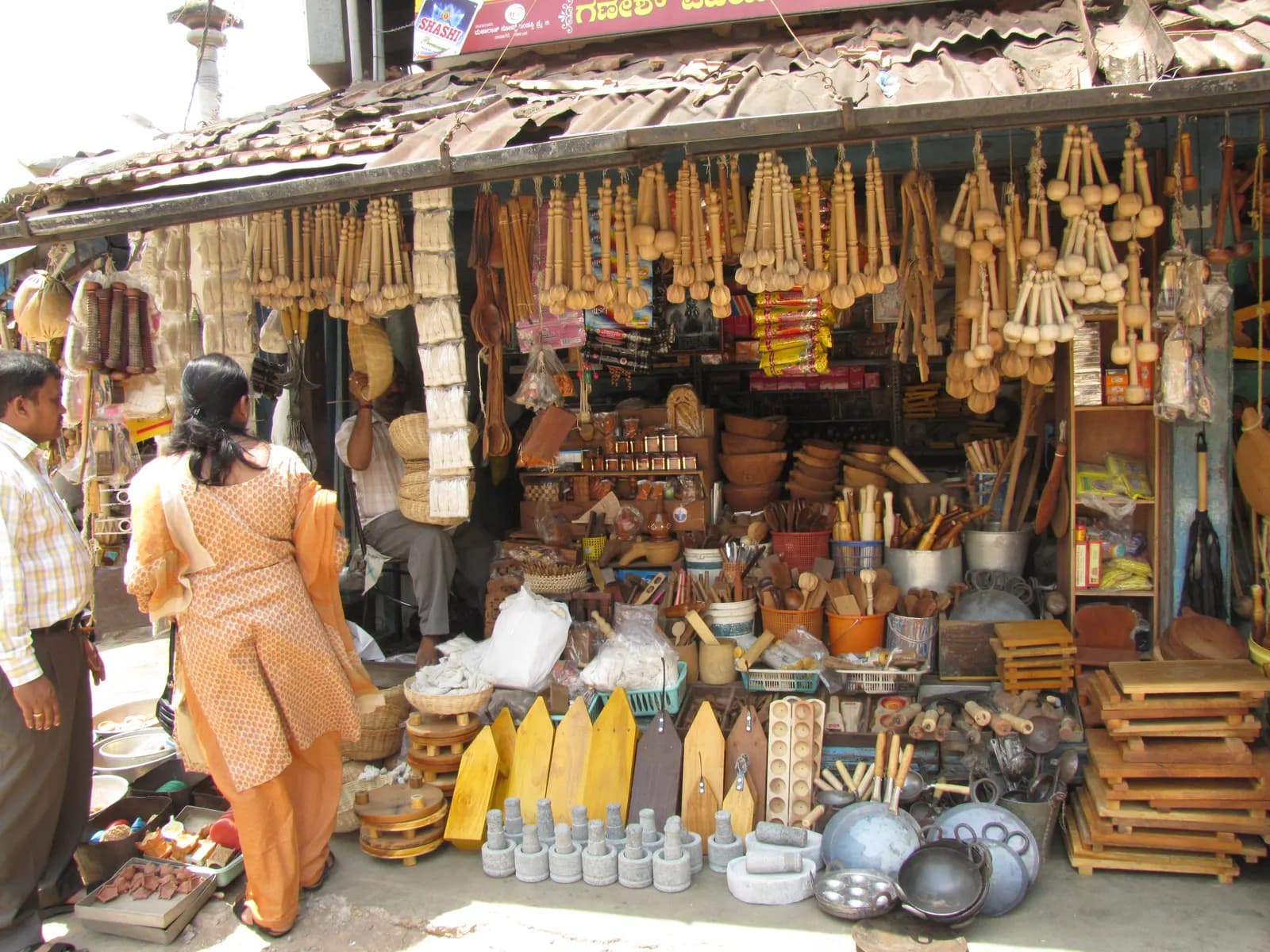 A handicraft and kitchenware shop on Car Street in Udupi, Karnataka, with wooden utensils hanging from the awning and stock spread on the pavement