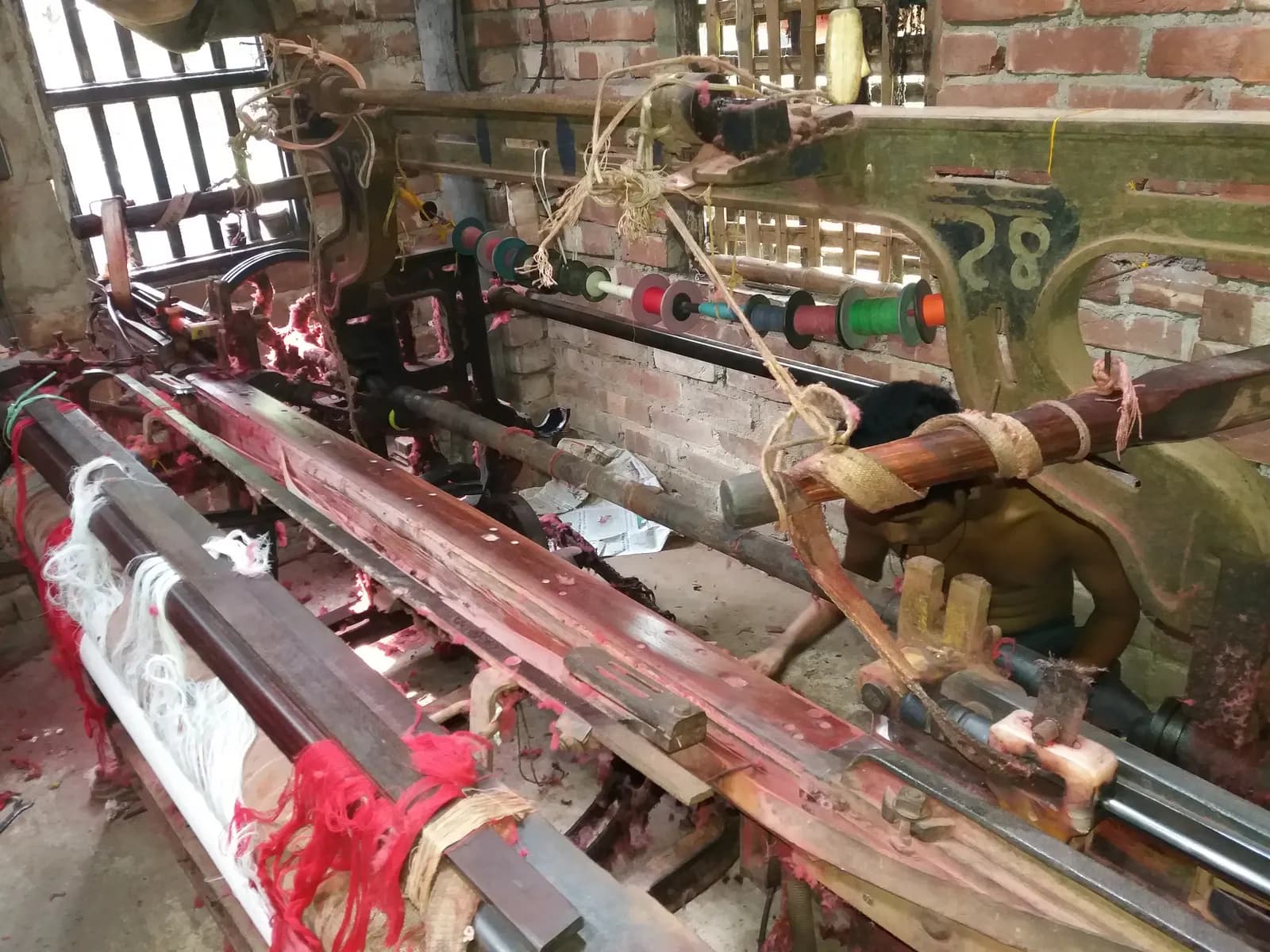 A worker adjusting a power loom with red and white yarn in a small brick-walled weaving shed in Hooghly district, West Bengal