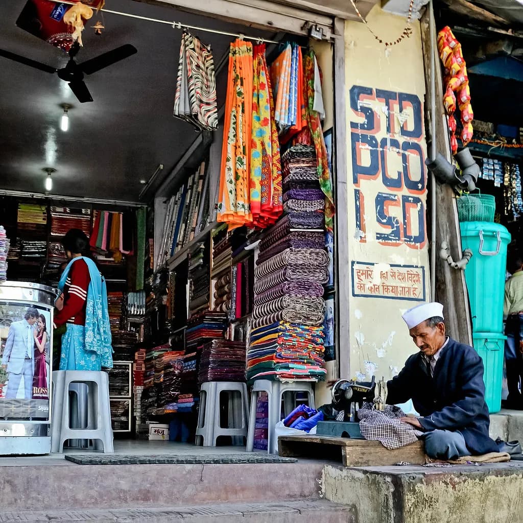 A cloth shop in India with stacked bolts of fabric on shelves and a tailor at a sewing machine outside