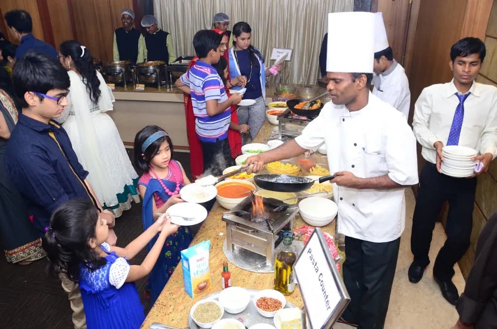 A chef running a live pasta counter at an Indian function buffet while staff in caps stand behind a row of chafing dishes