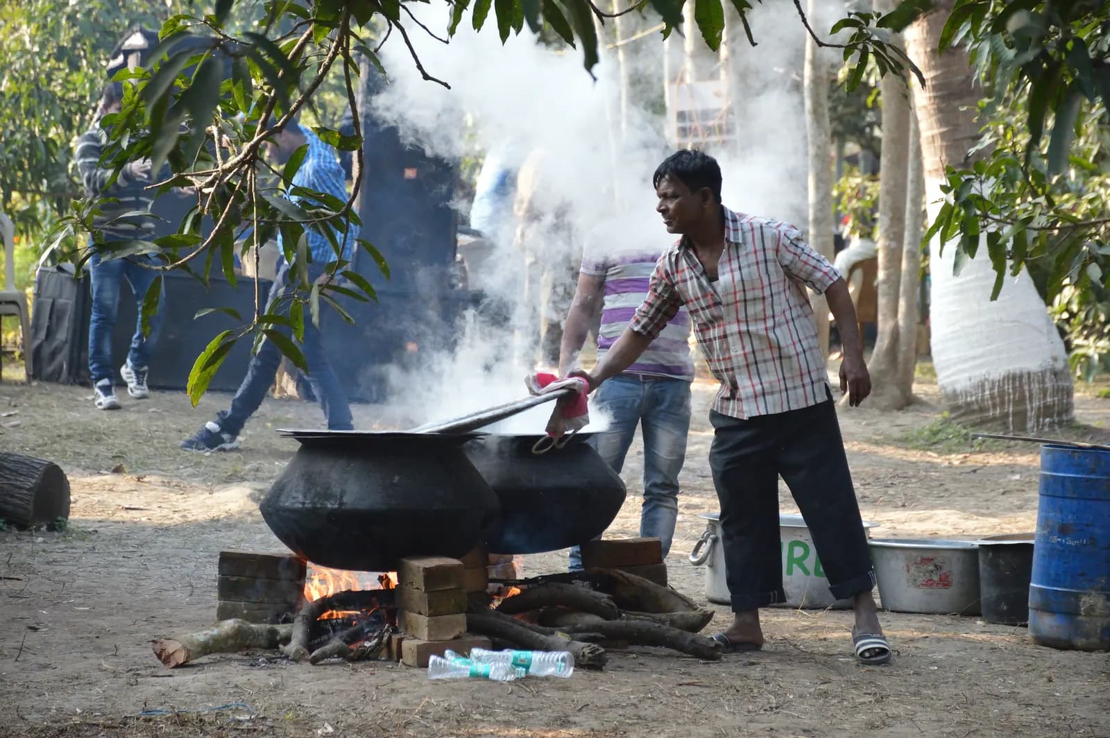 A cook lifting the lid on large handis set over a wood fire at an outdoor event site in North 24 Parganas, West Bengal