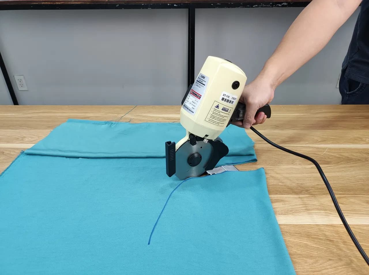 A hand-held round-knife fabric cutting machine slicing through a layer of blue knitted fabric on a cutting table