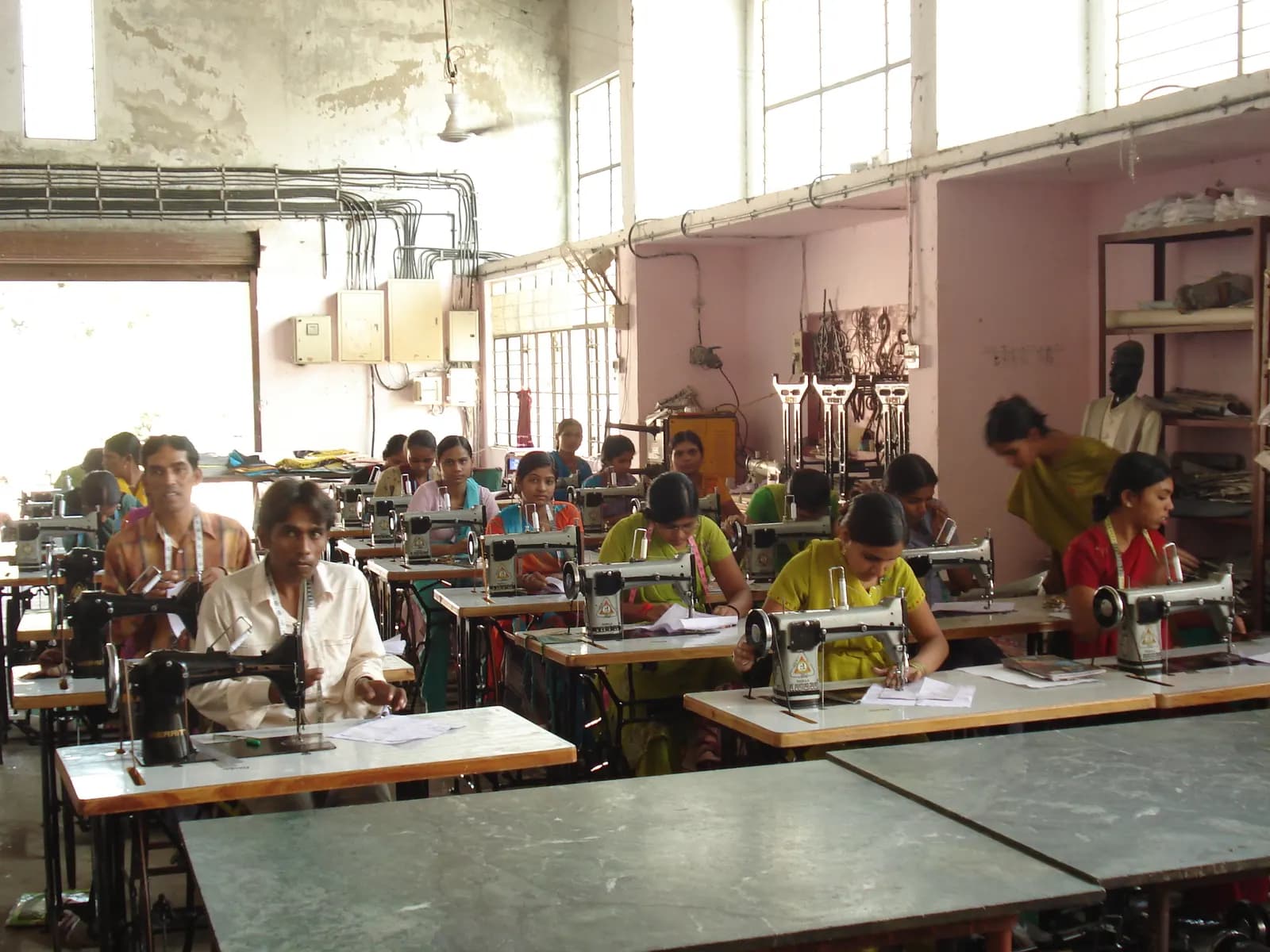 Rows of women operating single-needle sewing machines on the floor of a small garment unit in India