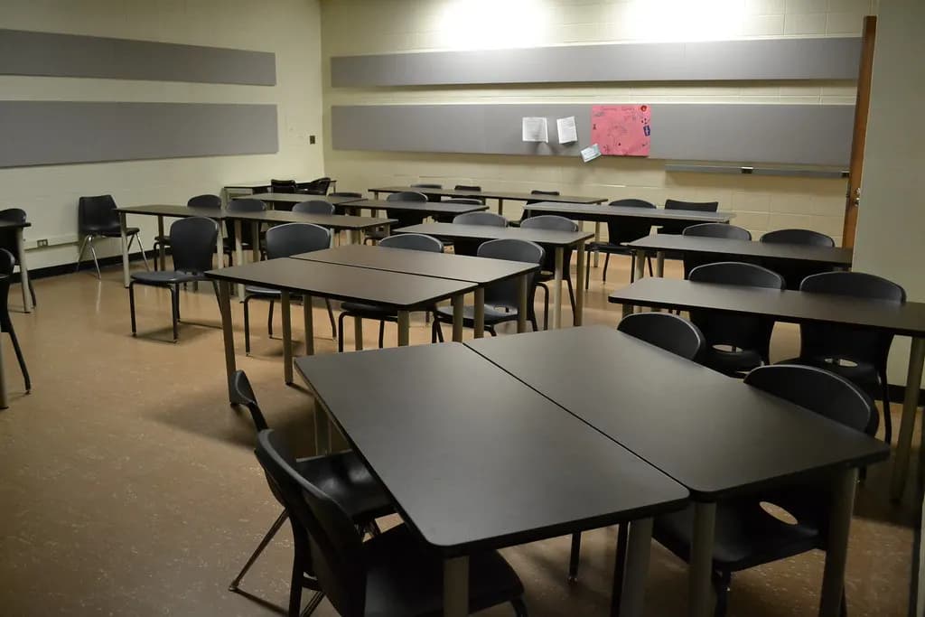 An empty classroom with grey tables pushed together in groups, black chairs and a long whiteboard along the wall
