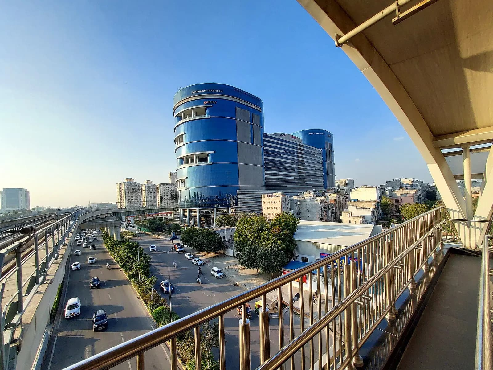 A curved blue glass corporate office tower beside a busy road in Gurugram, seen from a metro walkway