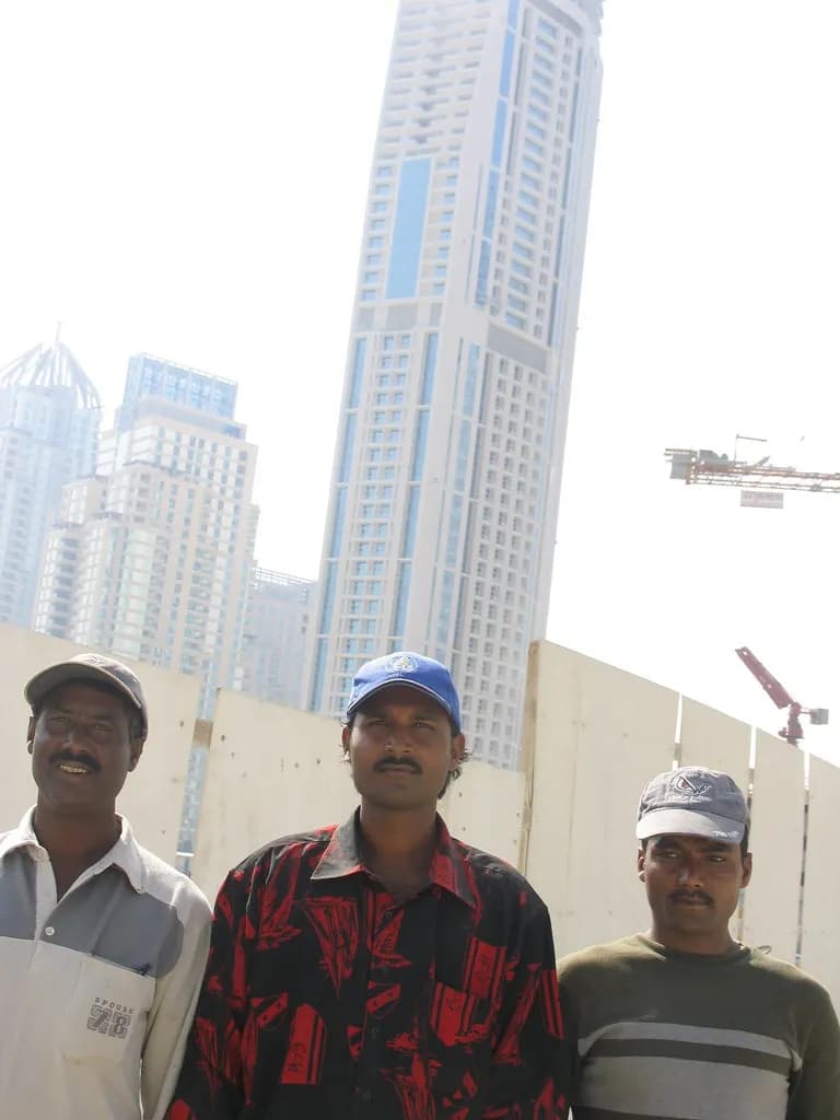 Three construction workers in caps standing at a building site with high-rise towers and a tower crane behind them