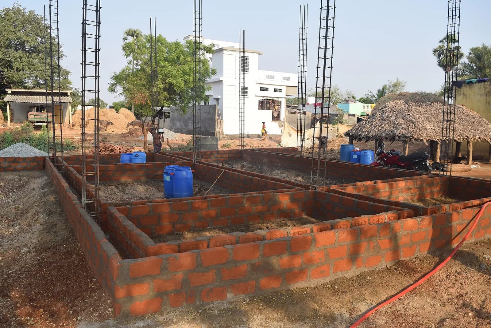 A house at plinth stage in India: brick plinth walls laid out in rooms, with column reinforcement bars standing ready for the next pour