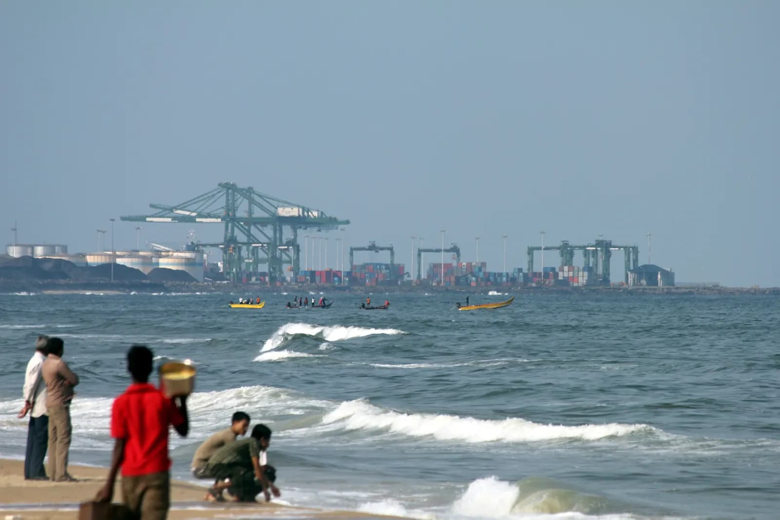 Gantry cranes and container stacks at Chennai Port seen across the sea from the beach, with people on the shore in the foreground