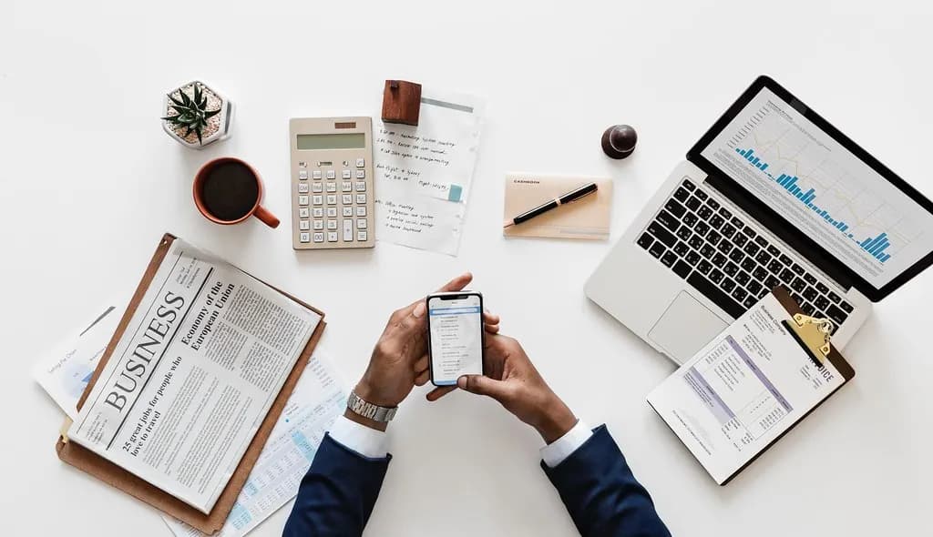 Top view of hands holding a smartphone over a desk with a laptop, calculator and business papers