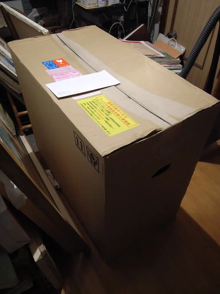 A large cardboard parcel with a shipping label and documents taped to the top, standing in a room
