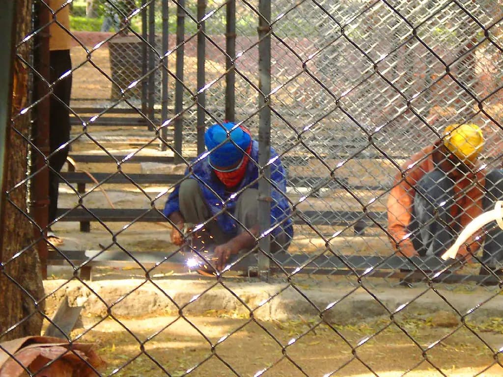 Two workers welding steel frames on the ground at a small fabrication job in India, seen through a chain-link fence