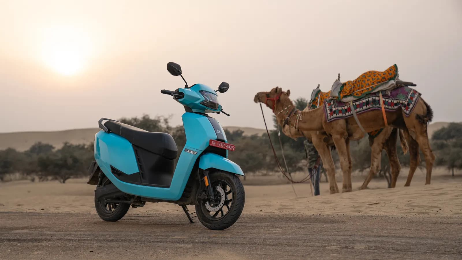 A light-blue electric scooter parked on a desert road in Rajasthan with camels in the background