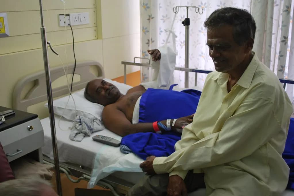 A patient with an injured arm resting in a hospital bed in India while a friend sits beside him