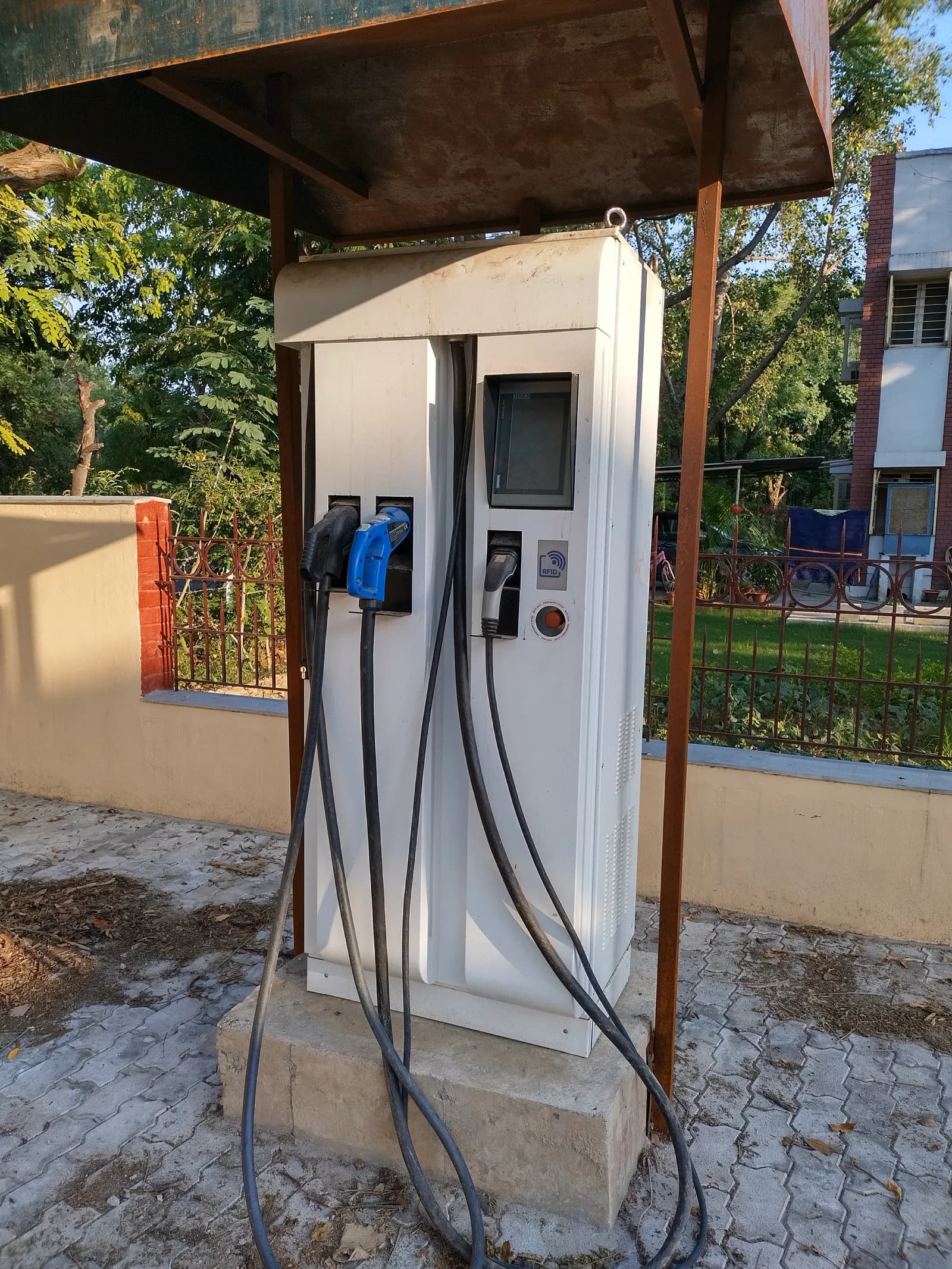 A free-standing DC fast charger with three charging guns and a touch screen under a small shelter on a campus in Kanpur