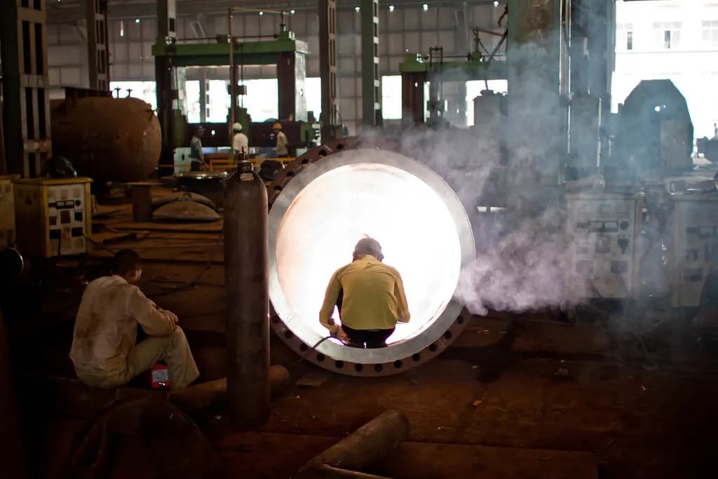 A welder working inside a large steel pipe section in an Indian heavy fabrication factory, with machines and workers behind