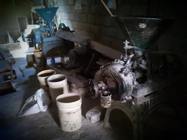 Inside a small Indian flour mill: grinding machines with blue hoppers, steel buckets of flour and a single motor driving the machines by belt