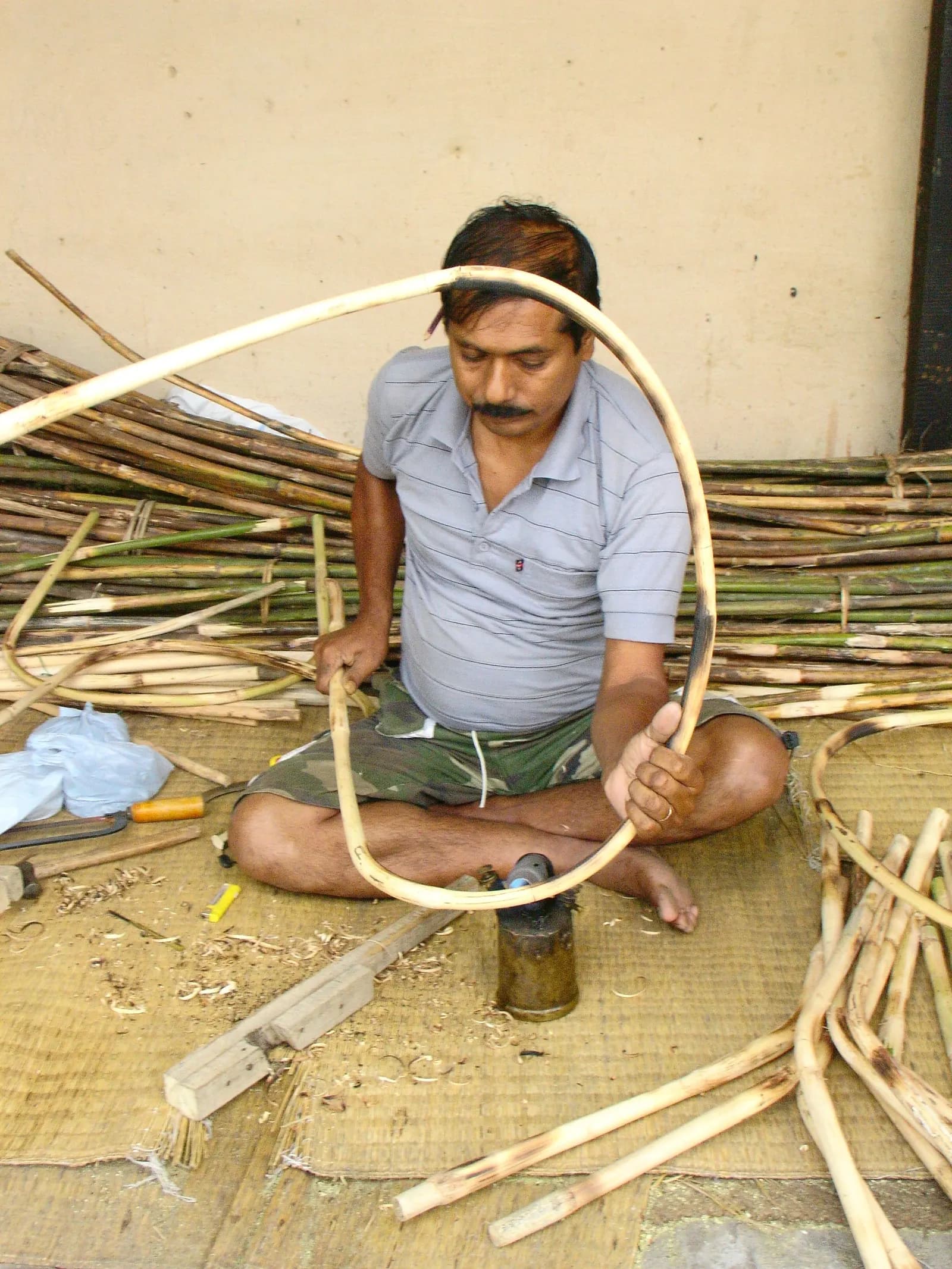 An artisan sitting on a mat bending a cane strip over a small burner to shape cane furniture, with bundles of raw cane behind him