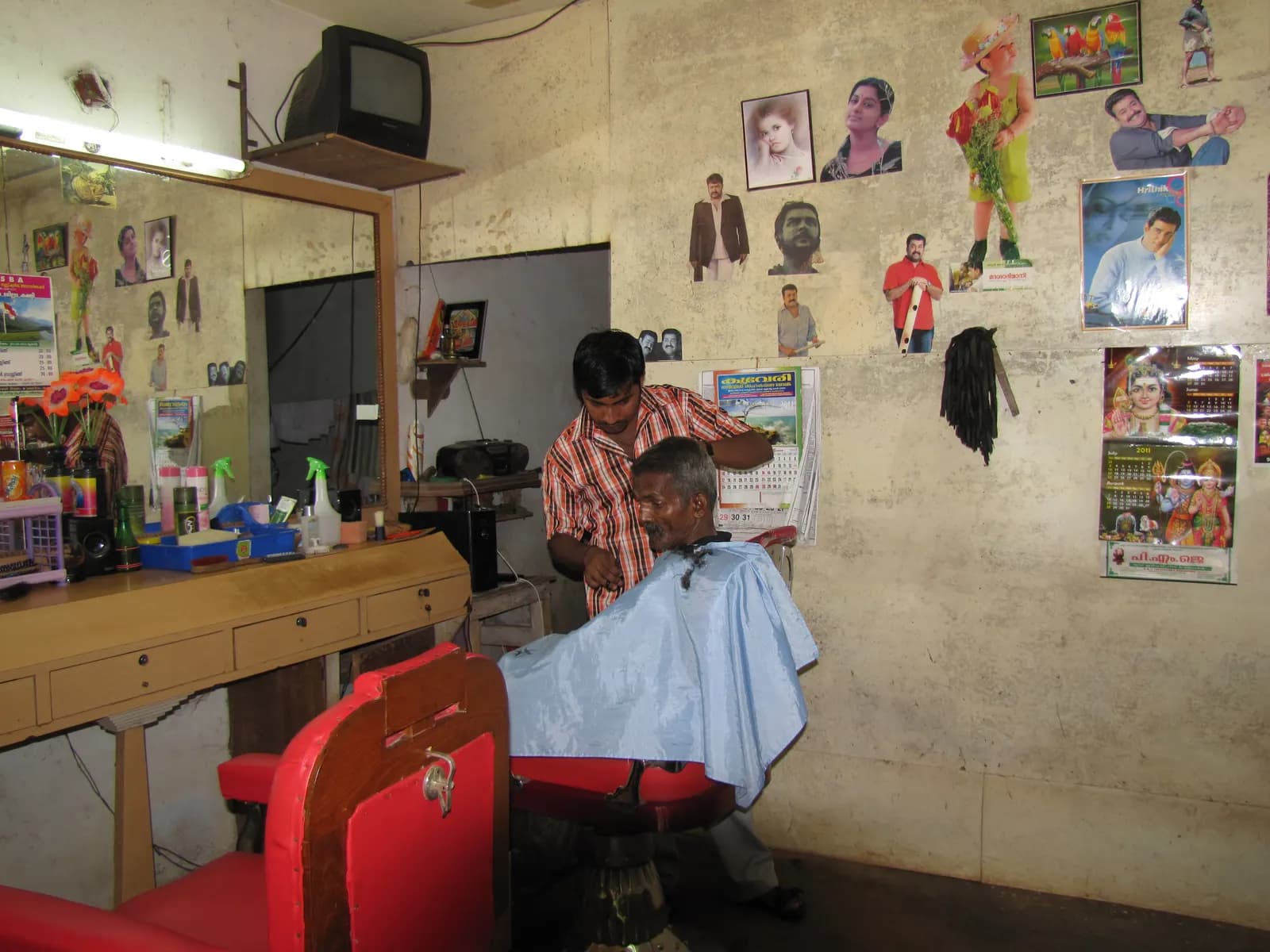 A barber giving a haircut in a small neighbourhood barber shop with a mirror, counter and posters on the wall