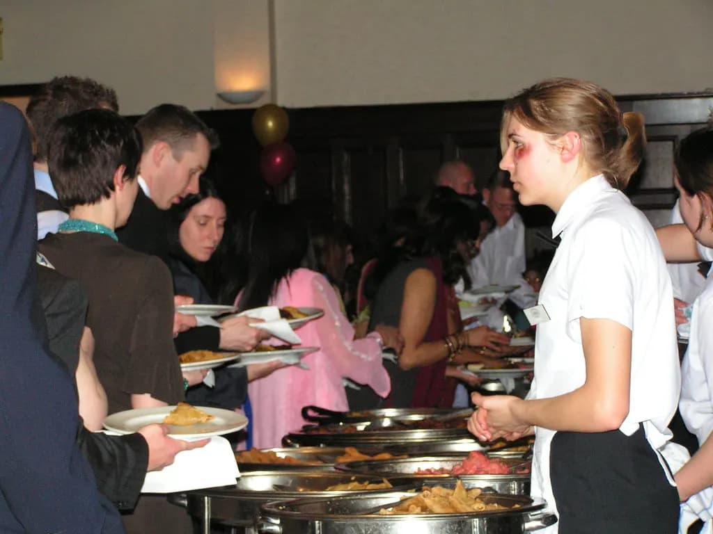 Guests and serving staff at a buffet counter lined with steel chafing dishes