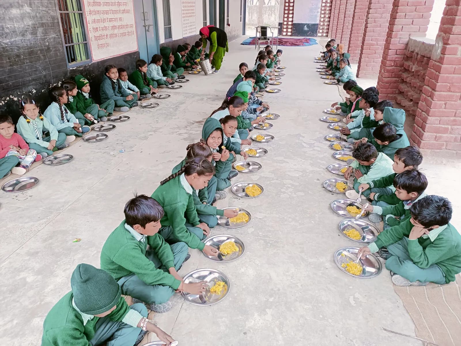 Schoolchildren in green uniforms seated in rows on a corridor floor eating a mid-day meal from steel plates