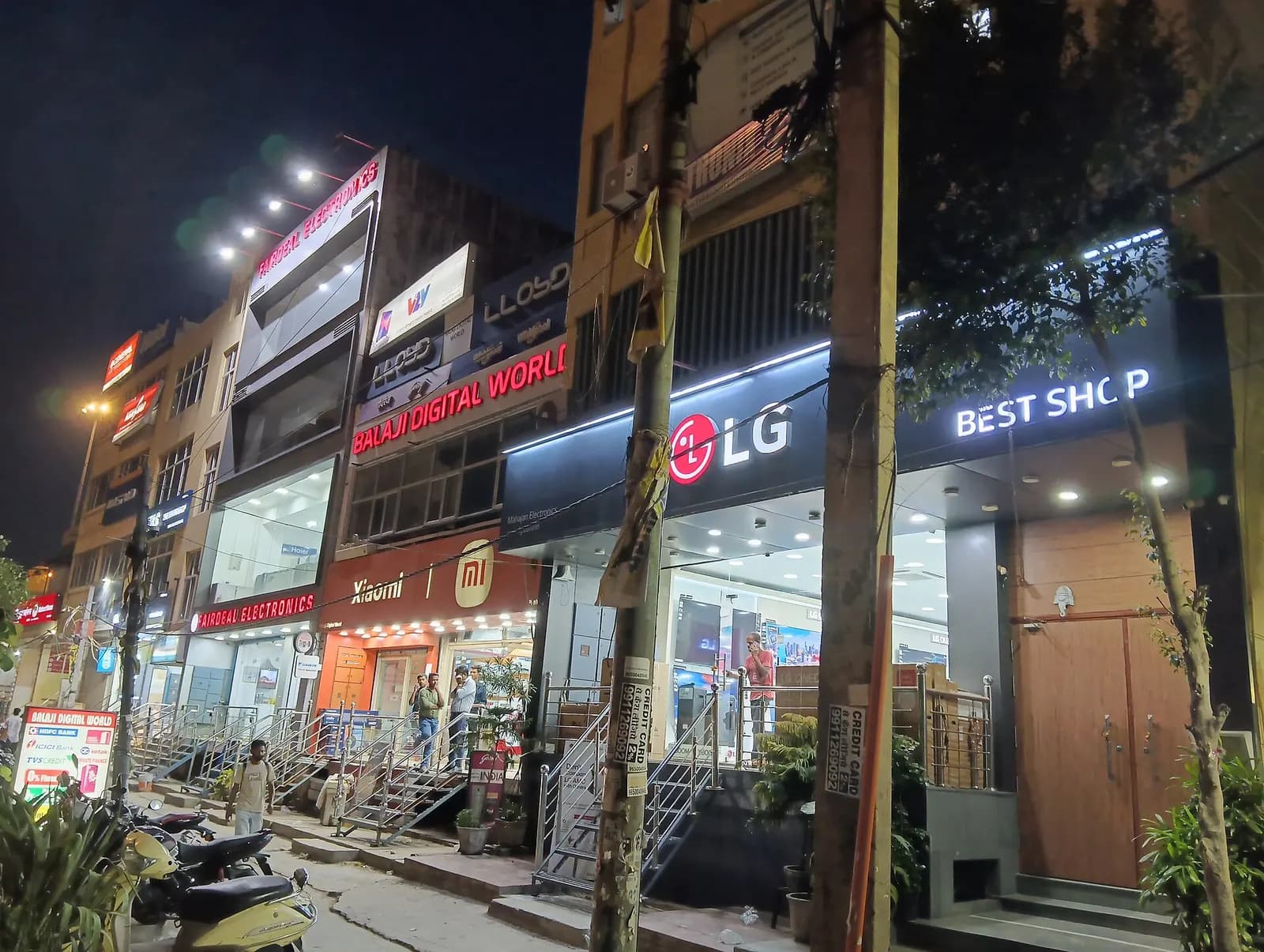 Home-appliance and electronics showrooms lit up at night on a market road in Dabri, Delhi