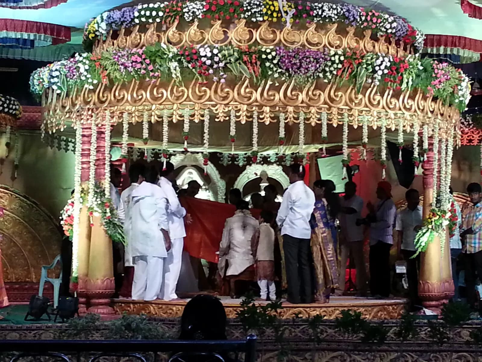 A wedding mandap decorated with flowers and a carved canopy, with guests standing on the stage