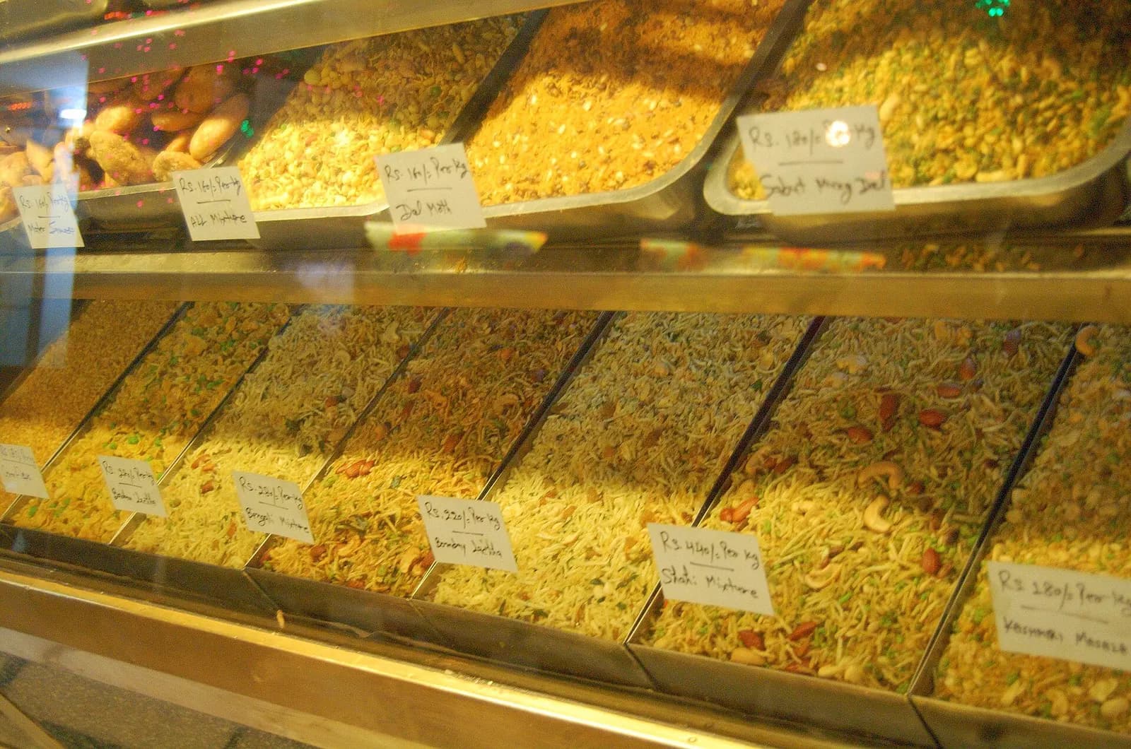 Trays of loose namkeen mixtures with handwritten price-per-kg labels in a sweet shop display counter