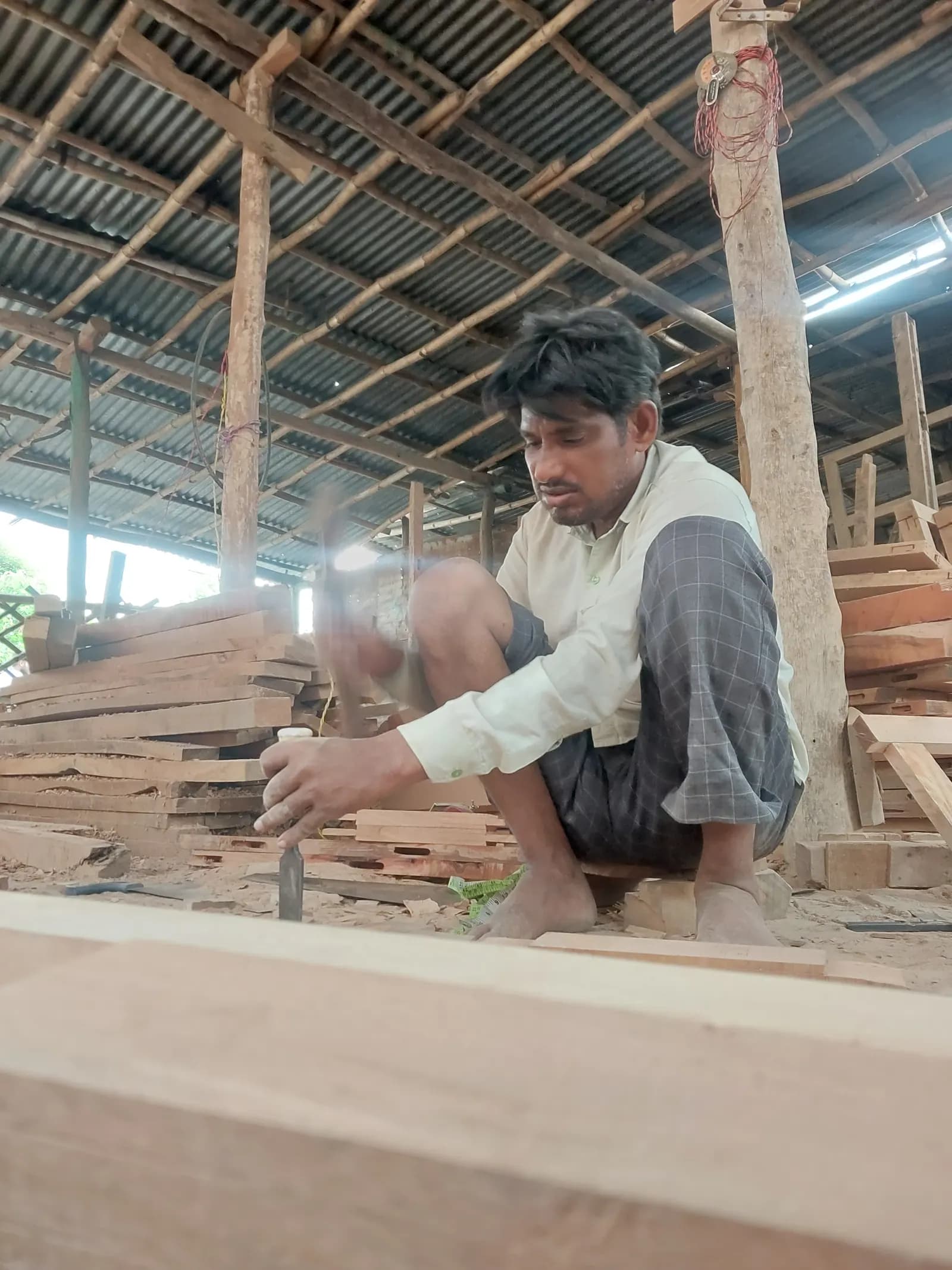 A carpenter chiselling a wooden plank in a timber workshop with stacked wood