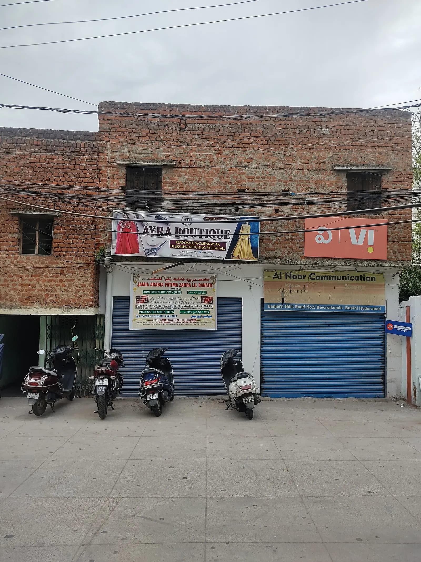 A row of small ground-floor shop units with shutters and signboards, with scooters parked in front