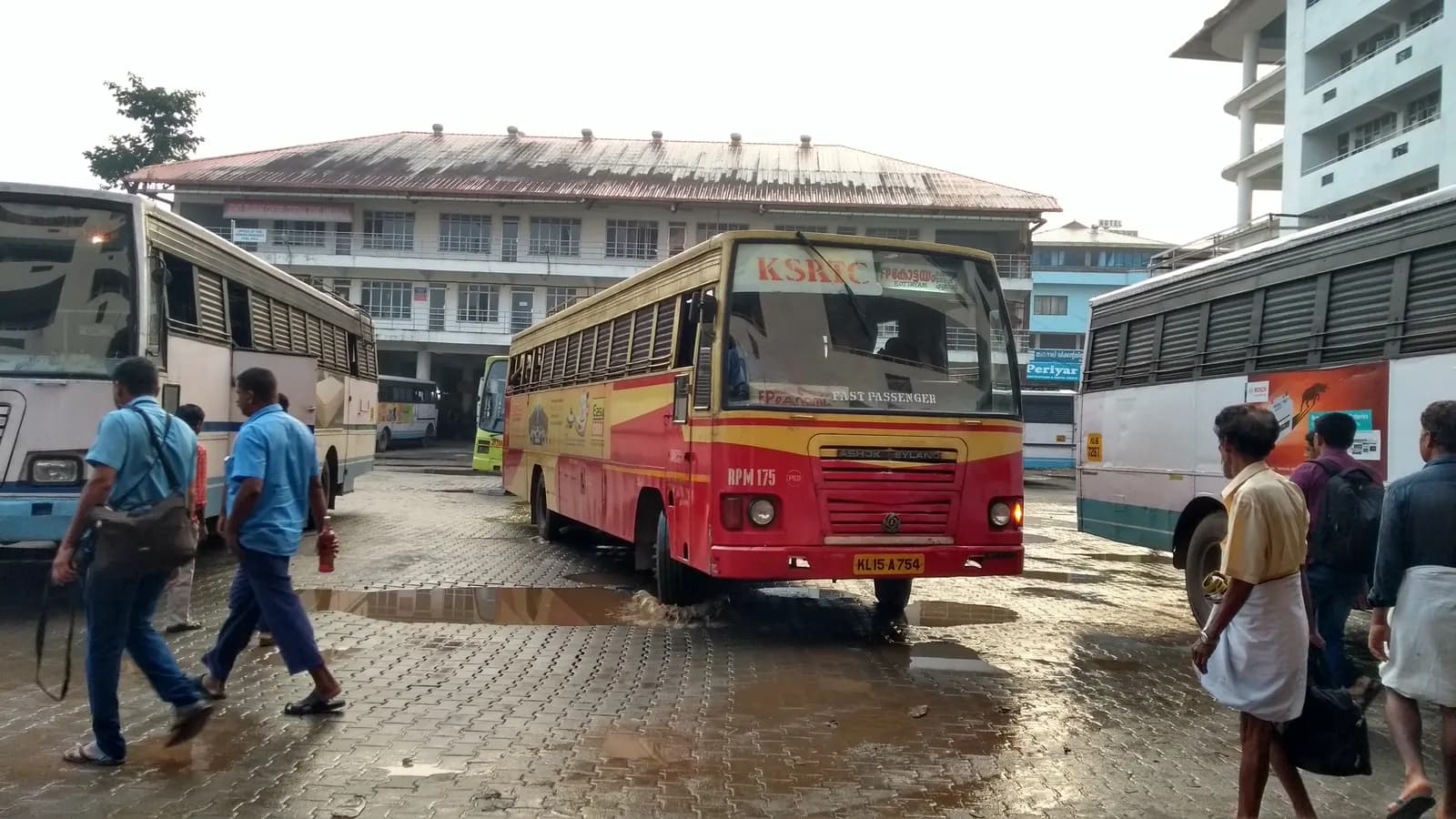 A red and yellow non-AC state transport bus pulling into a bus station in Kerala, with passengers walking nearby