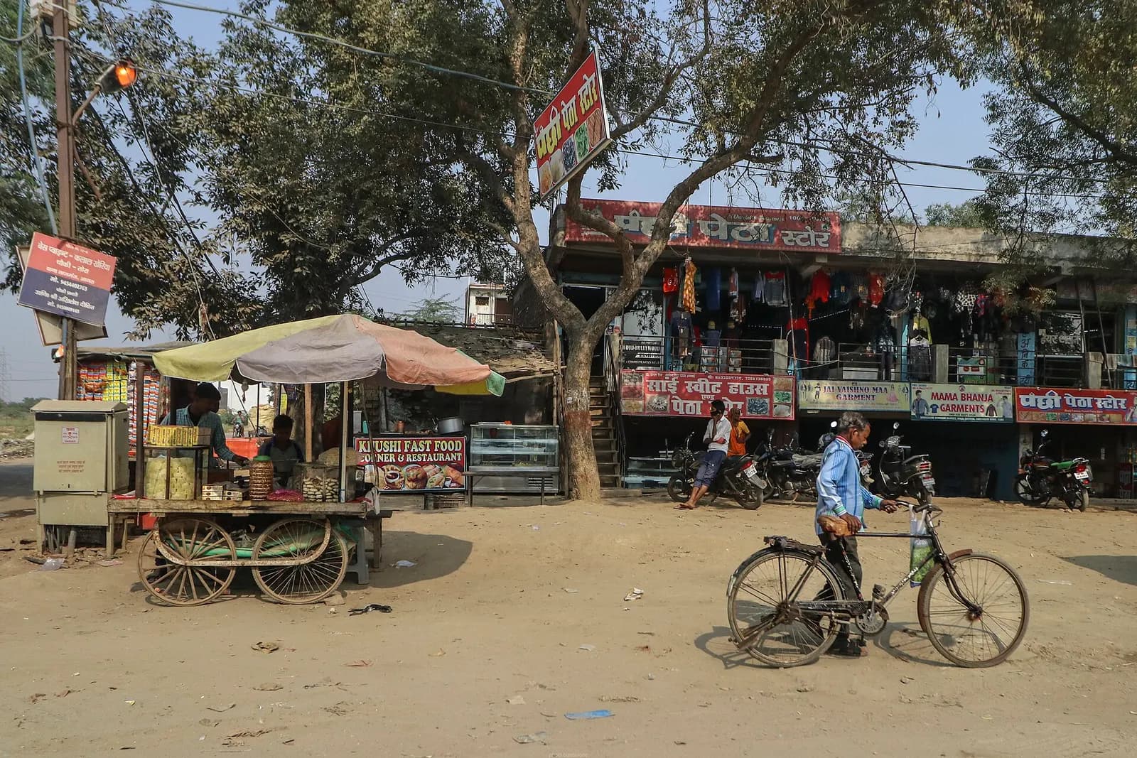 A man wheeling a black roadster bicycle past roadside shops and a food cart in Uttar Pradesh