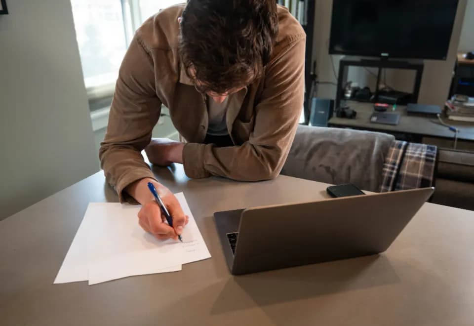 A man at a kitchen table writing notes on paper next to an open laptop