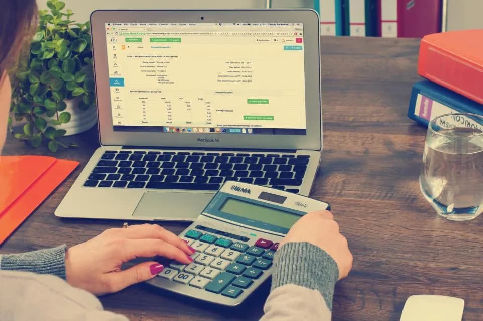 Hands working a desk calculator in front of a laptop showing an accounting spreadsheet