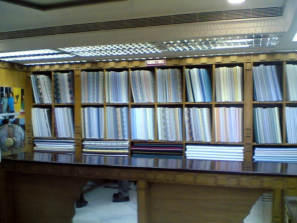 Bolts of fabric stacked on wooden shelves behind the counter of a small cloth shop in India