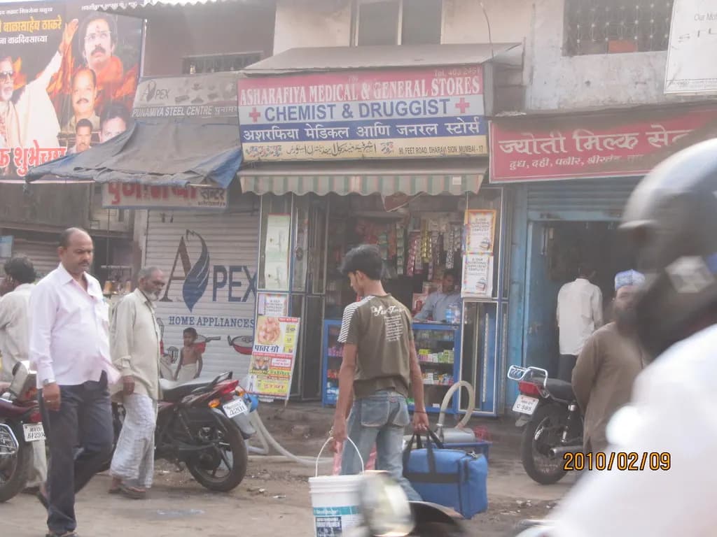 Row of small roadside shops, including a chemist and a general store, on a busy street in Dharavi, Mumbai
