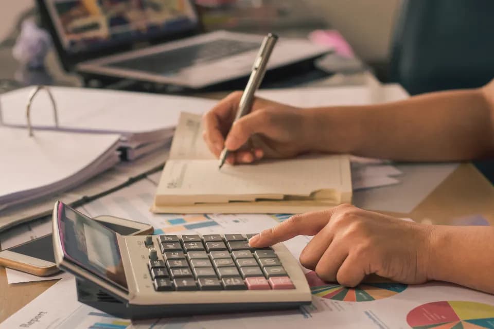 A person writing in a notebook beside a calculator, ring binders and printed charts