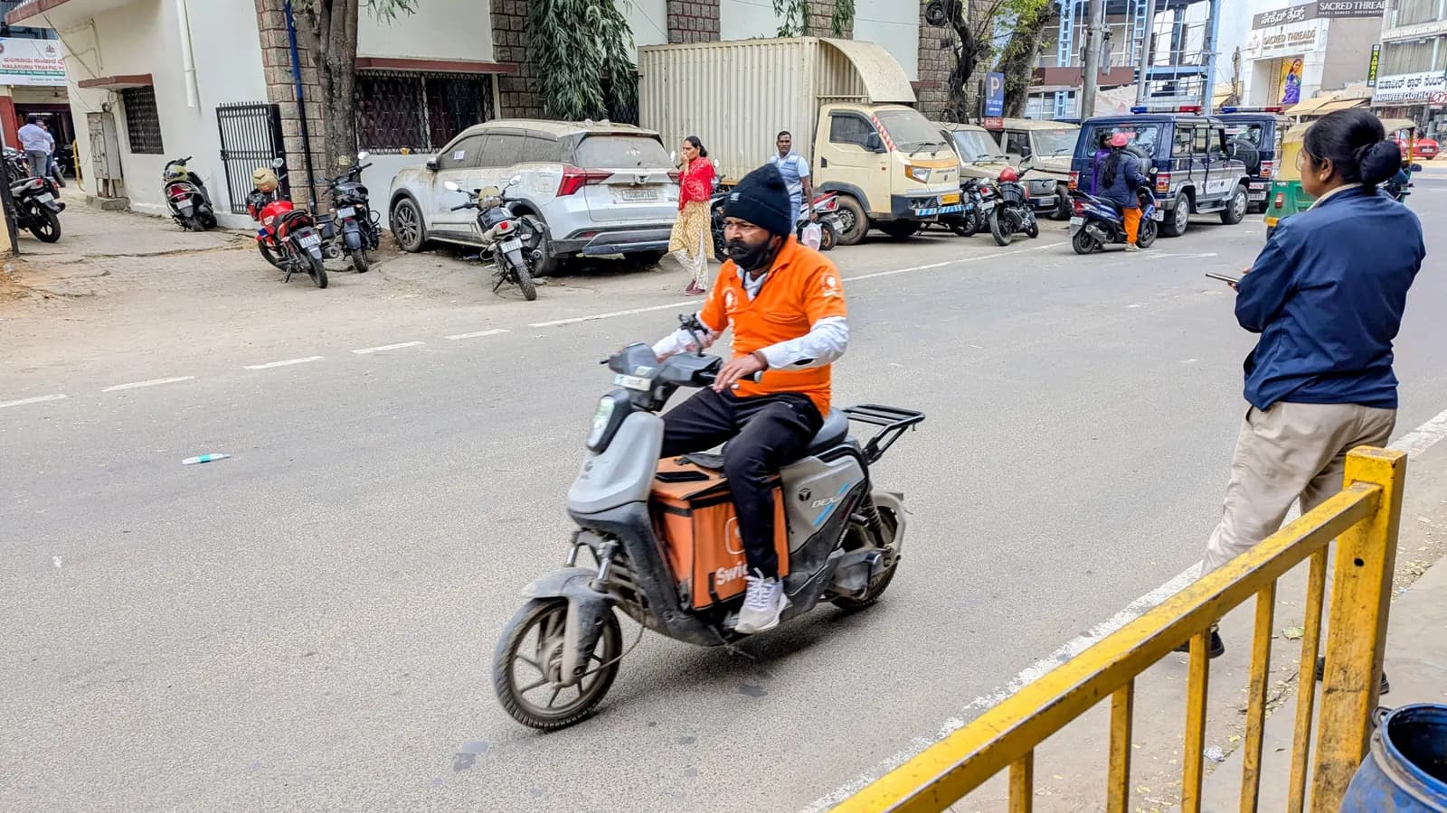 A food delivery rider in an orange jacket with a delivery box on an electric scooter on an Indian street