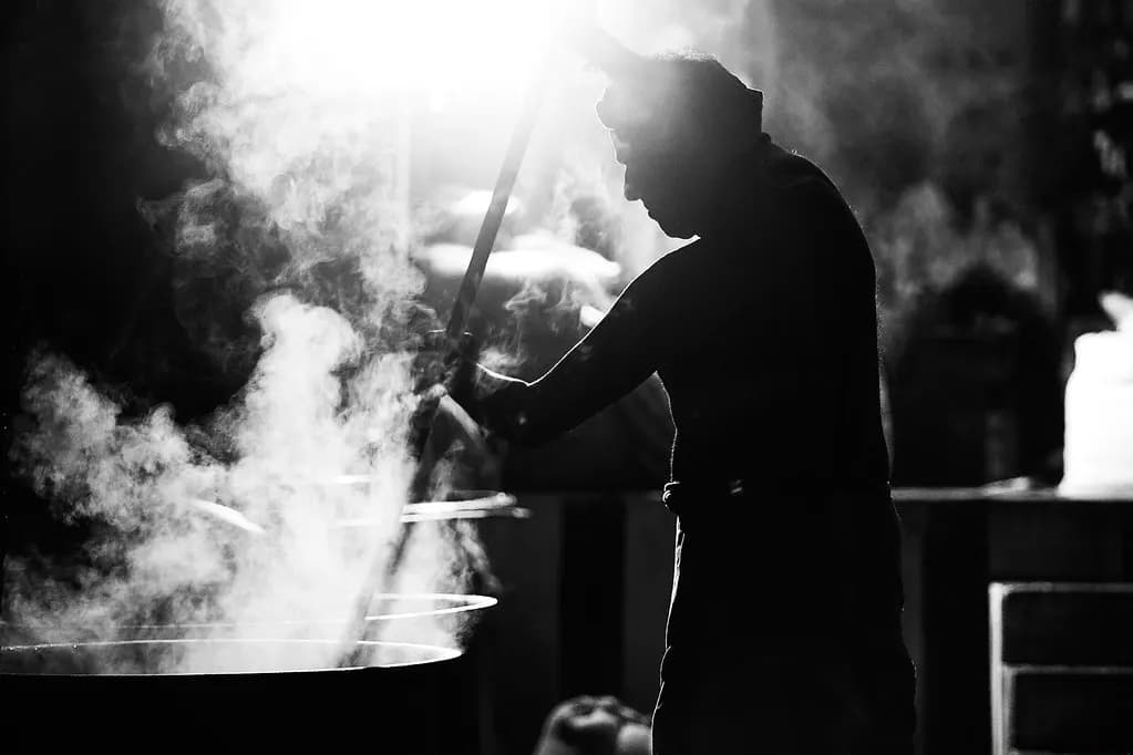 A cook stirring a large steaming pot with a long ladle in a commercial kitchen