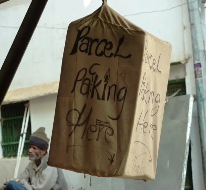 A cloth-wrapped parcel hanging outside a small parcel-packing shop in Dharamsala, with a hand-written sign reading Parcel Packing Here